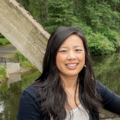 A photo of Dr. Jamie Cho. She's Asian, brunette, with long straight hair. She's smiling in the photo, and appears to be standing in front of a river. She's wearing a blue cardigan and white shirt.