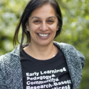 A photo of Ninderjit Gil. She is smiling, her hair is tied back, and she's wearing a black shirt under a gray cardigan. Her shirt says "Early Learning & Pedagogy & Community & Research-Based...". The background appears to be blurred bushes or trees.