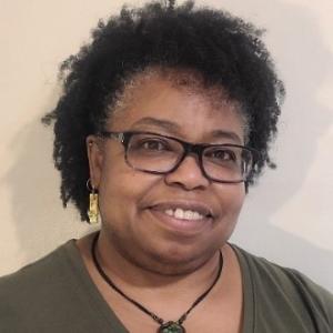 A photo of Dr. Cynthia. She is a black woman with short, coily black hair. She's smiling, wearing black glasses, a black necklace, and green v-neck shirt. She's also wearing gold earrings. 