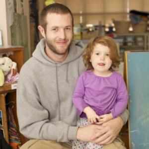 A white father sitting with his daughter on his lap. The father has short brown hair and is wearing a gray hoodie. The daughter is calm and is wearing a purple long sleeve shirt. Her hair is short and a bit red. 