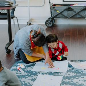 A photo of a caregiver playing with a toddler. They are sitting on the floor, doing some kind of art project together.