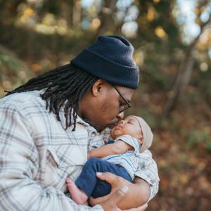 a black father kissing his newborn baby. He's cradling the baby in his arms. The father is wearing a flannel shirt, dreads, glasses, and a blue beanie. The baby is wearing a blue shirt and blue pants. They are standing in what appears to be a forest or park.