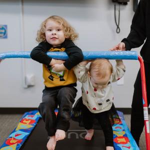 two white toddlers playing on a jumping mat. They have short blonde hair. Looks like they're siblings. The one on the left may be around 2 years old, and the one on the right probably 1 years old. An adult wearing all black is supervising them. 