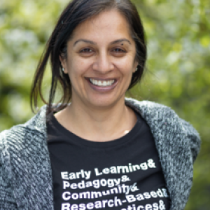 A photo of Ninderjit Gil. She is smiling, her hair is tied back, and she's wearing a black shirt under a gray cardigan. Her shirt says "Early Learning & Pedagogy & Community & Research-Based...". The background appears to be blurred bushes or trees.